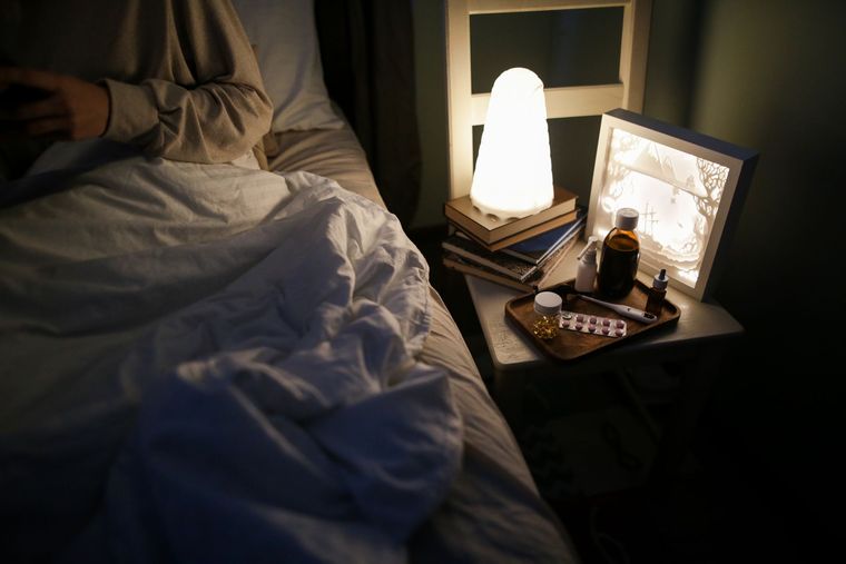 Table de nuit encombrée : lampe, livres, médicaments, près d'un lit défait